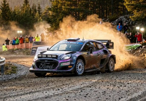 A multi-colored Ford rally car races along a gravel road, kicking up dust on a sunny day. People watch from the side of the track. The surrounding area is hilly with green trees.